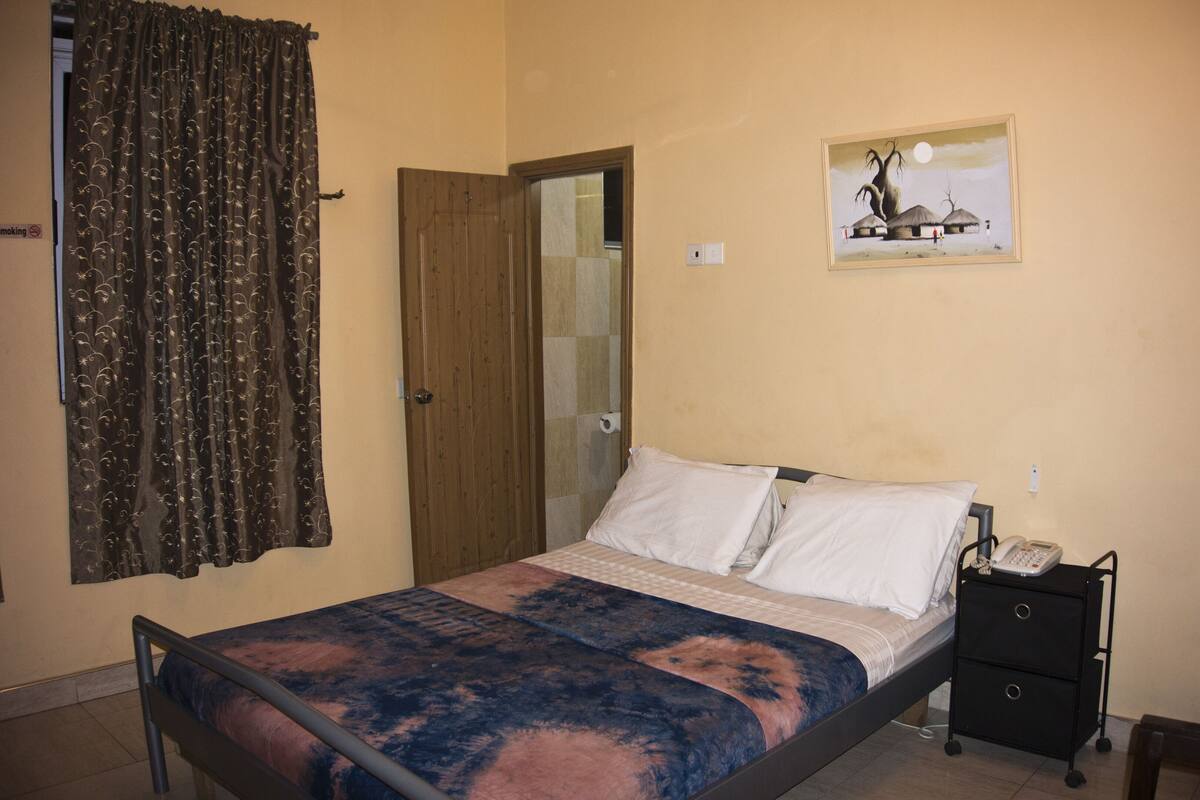 Quarto com cama, mesa de cabeceira com telefone, cadeira, quadro na parede e porta.