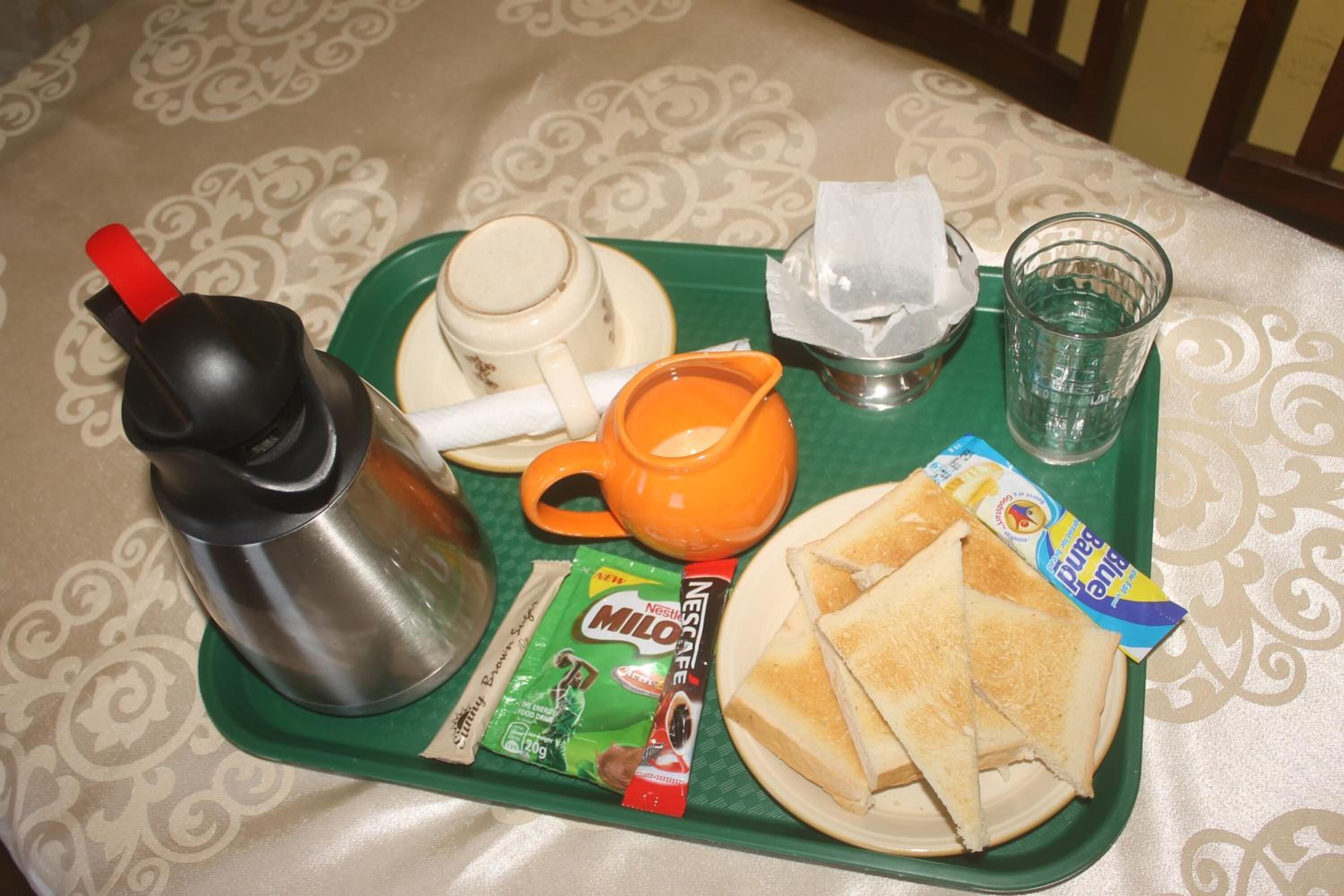 Uma bandeja com uma garrafa térmica, uma caneca, um pacote de Milo, um pacote de Nesca, um copo de água e um prato com torradas.