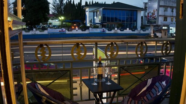 A balcony with two chairs and a table, overlooking a street with cars and buildings. - Jesiwa B's Little Palace (Accra)
