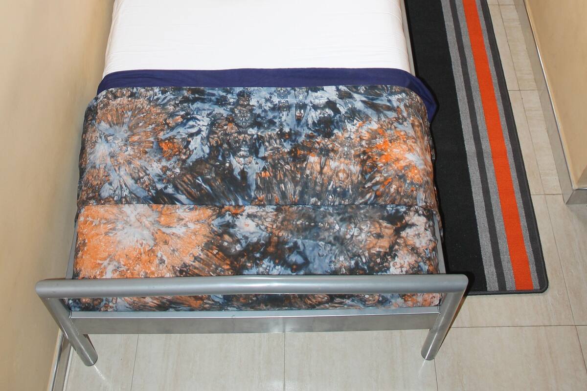 Uma cama bem arrumada com almofada estampada, mesa de cabeceira de madeira com abajur e tapete listrado sobre piso de azulejo.