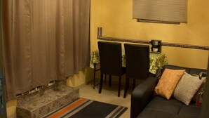 A small room with a dining table, two chairs, a sofa with cushions, a window with blinds, and a curtain. - Jesiwa B's Little Palace (Accra)