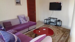 A living room with a sofa, a coffee table, a TV mounted on the wall, and a small table with a phone and a book. - Georgetown Court (Accra)