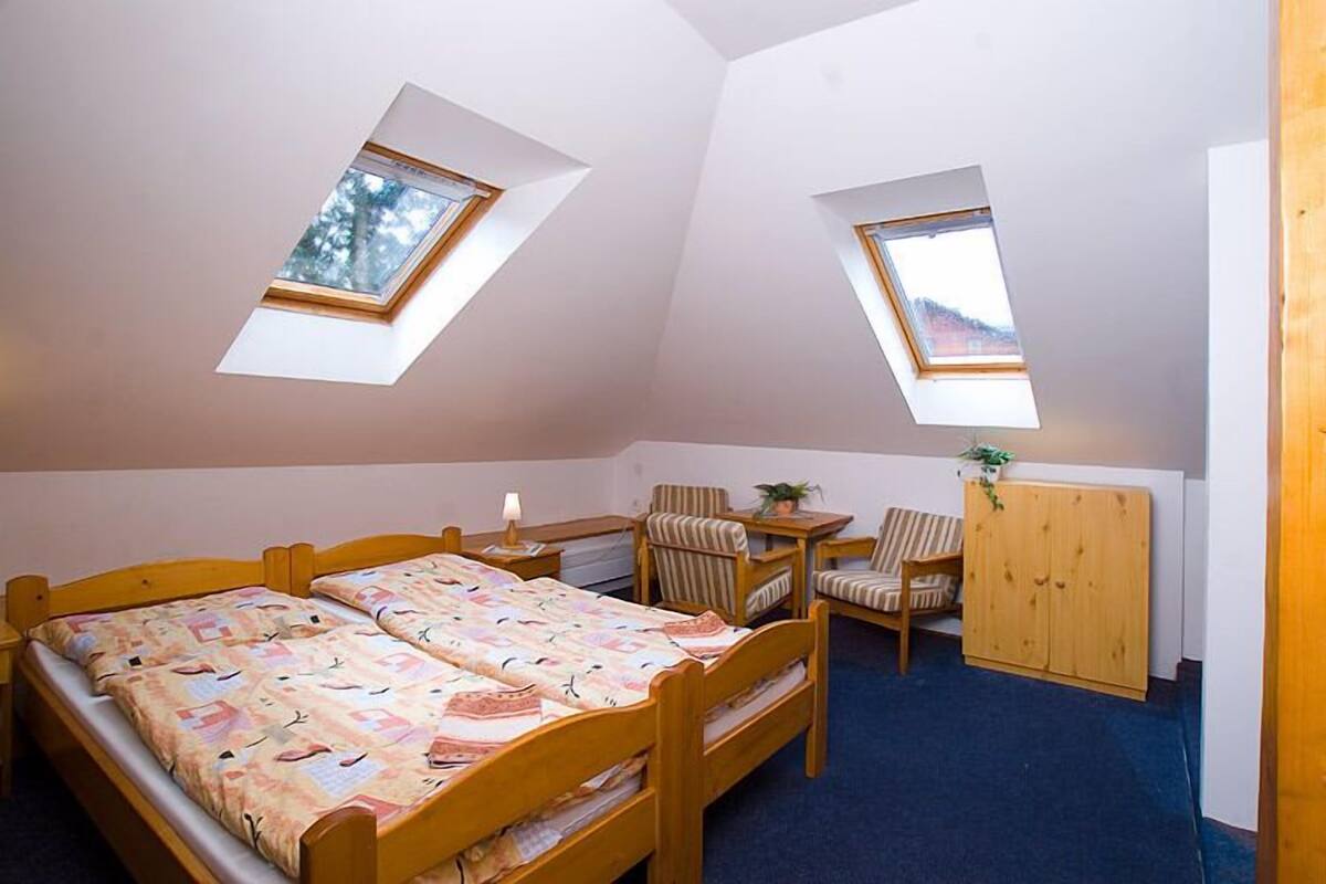 Quarto pequeno e acolhedor com teto inclinado, duas camas, uma escrivaninha, uma cadeira e um guarda-roupa.