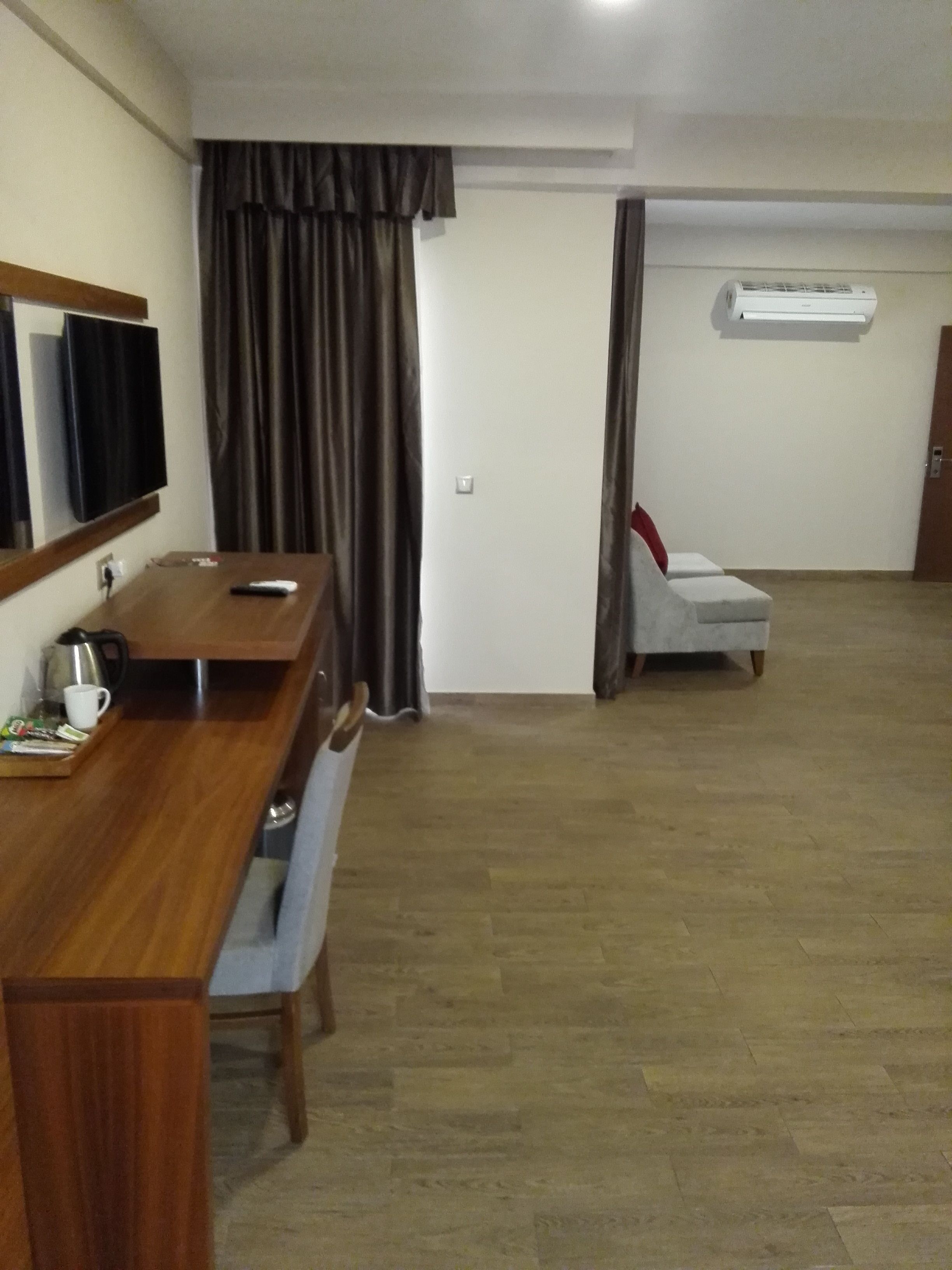 A hotel room with a wooden desk, a television, a chair, a small table with a cup and remote, and an air conditioning unit on the wall.