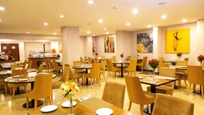 Uma sala de jantar ampla, com várias mesas e cadeiras, disposta em uma configuração moderna com um ponto central de serviço.