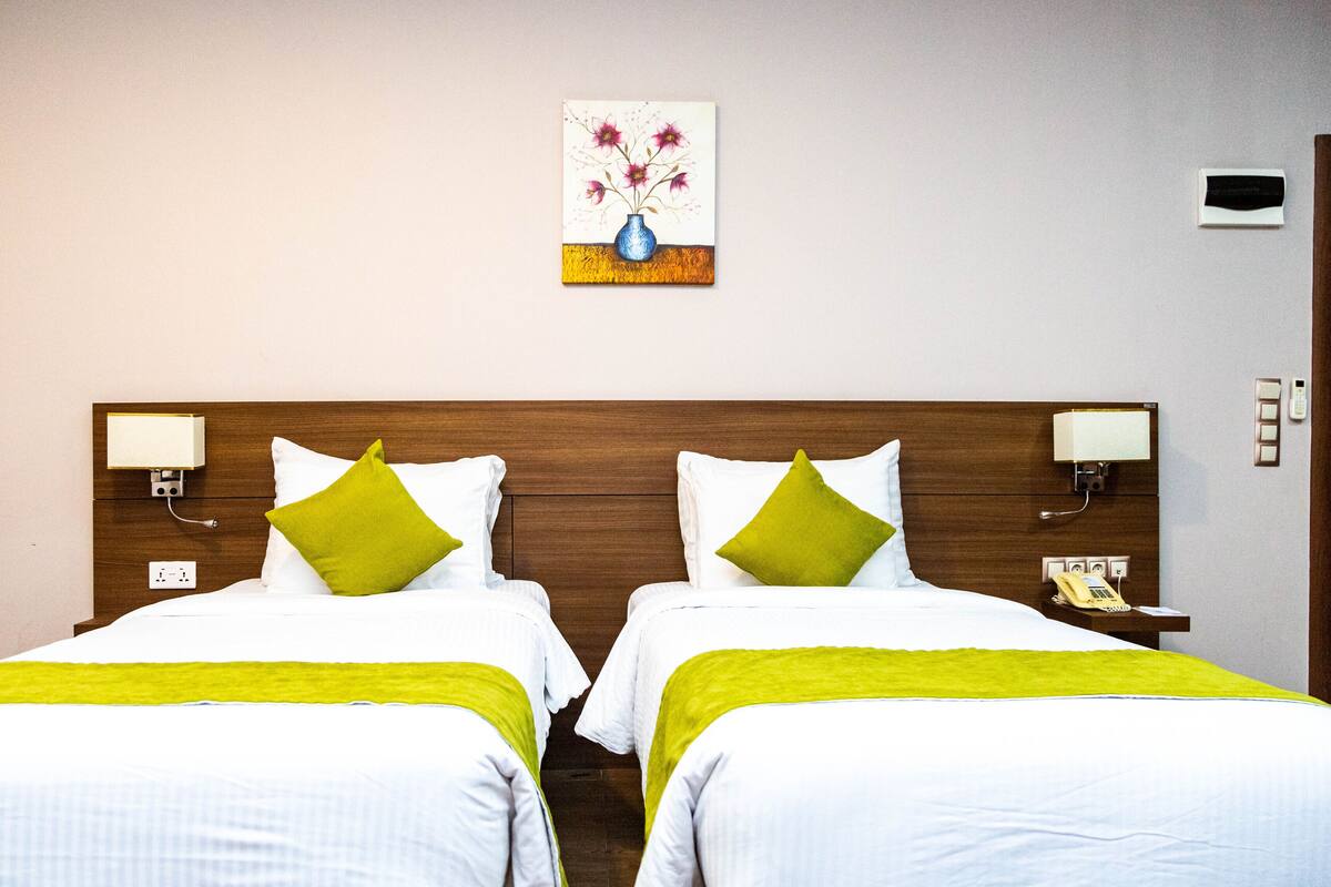 A hotel room with two beds, each with white bedding and green pillows, a wooden headboard, bedside lamps, a painting of flowers, and a telephone on the nightstand.