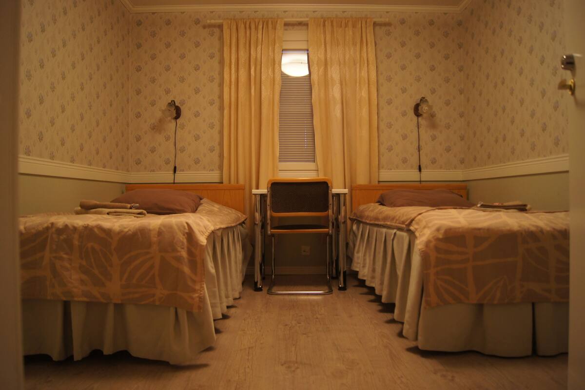 Quarto com duas camas, uma mesinha e uma janela com cortinas.