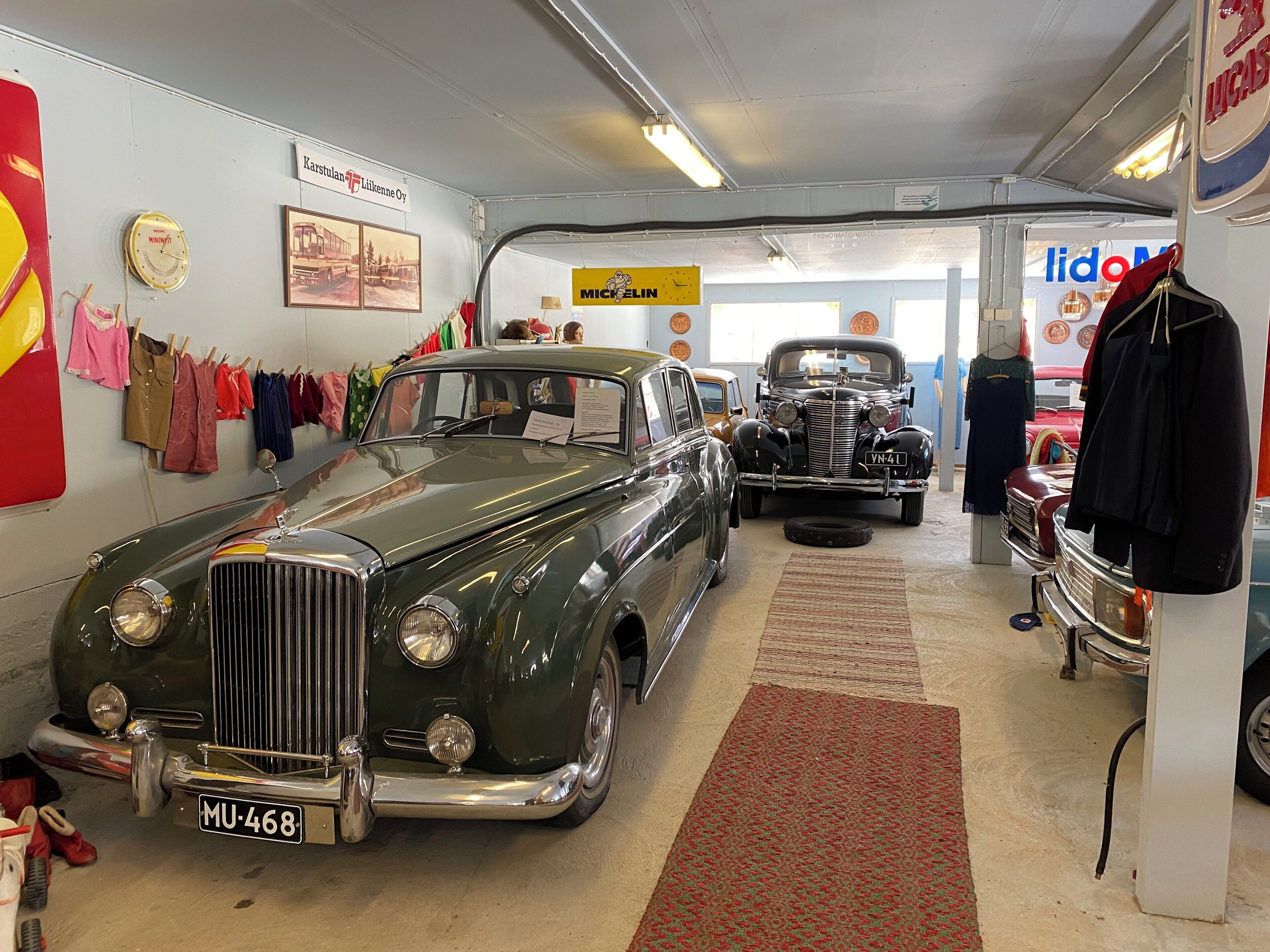 A vintage car showroom with classic cars on display, a clock, and various items on the walls.