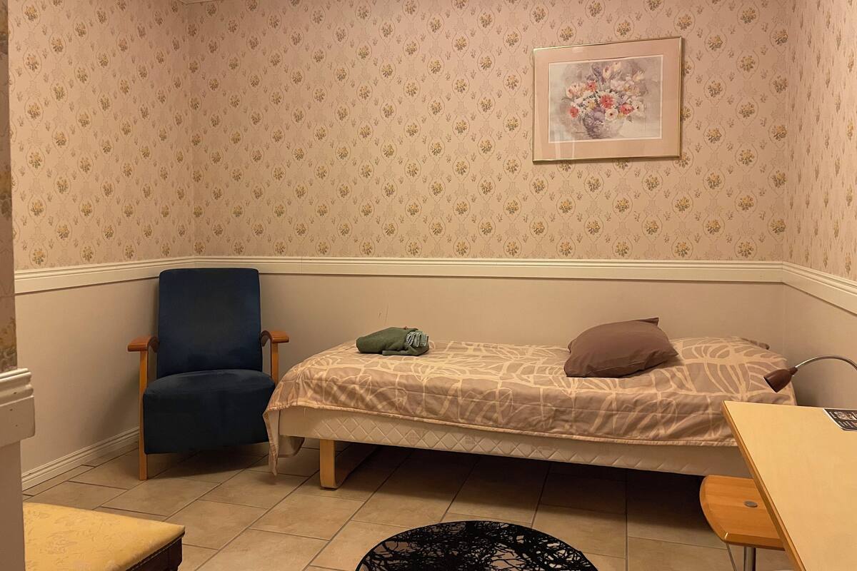 Um quarto pequeno e acolhedor com uma cama de solteiro, uma cadeira, uma mesa e um quadro na parede.