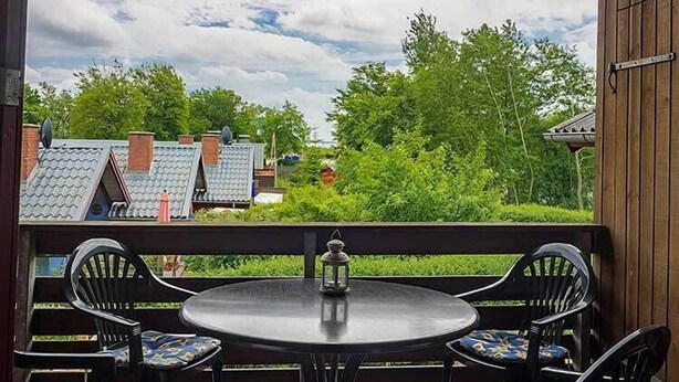 A covered outdoor seating area with a round table and four chairs, a lantern, and a view of houses and trees under a cloudy sky. - Ferienwohnung Polly (Gluecksburg)