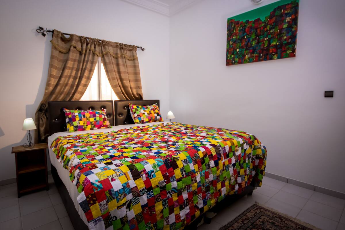 Ein Schlafzimmer mit einem großen Bett, einem Nachttisch, einer Lampe, einem Fenster mit Vorhängen und einem farbenfrohen Bild an der Wand.