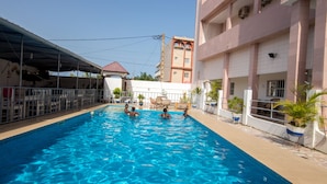 A swimming pool with clear blue water, surrounded by a covered area and a building with multiple balconies. - Jeane's Appart Hotel (Lomé)