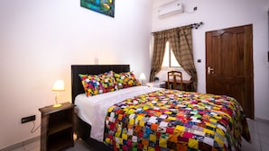 A bedroom with a bed, a nightstand, a window with curtains, a door, and a wall-mounted air conditioner. - Jeane's Appart Hotel (Lomé)