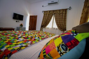 A bedroom with a colorful quilted bedspread, a flat-screen TV mounted on the wall, and a window with curtains.