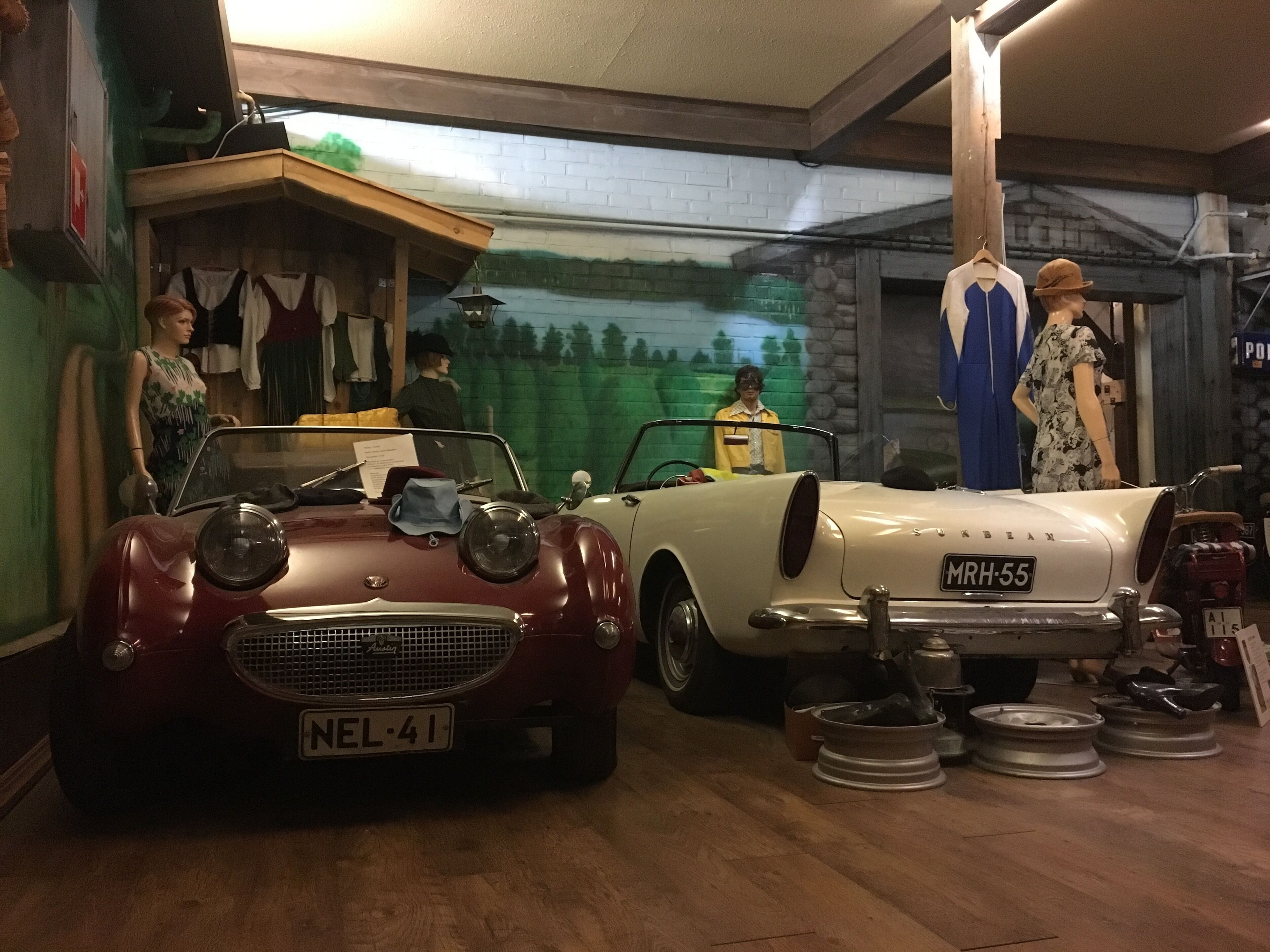 A vintage car showroom with two classic cars, a mannequin dressed in a hat, and a mural on the wall.