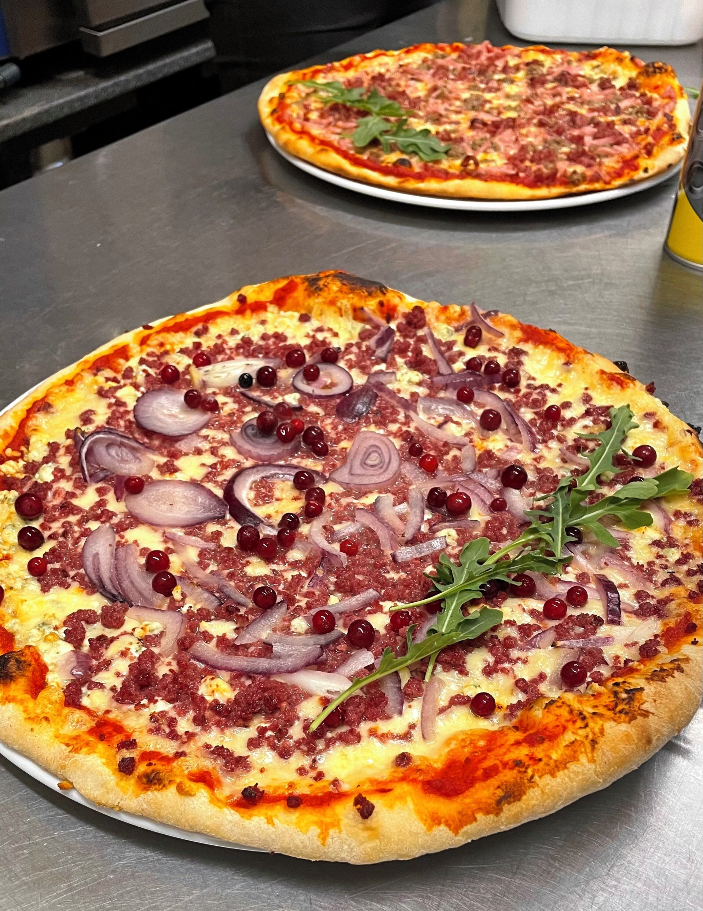 Two pizzas on metal trays with toppings like onions, arugula, and red berries.
