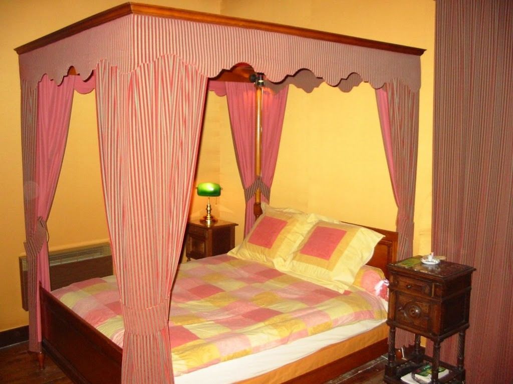 Una cama con dosel y cortinas, con un edredón acolchado, una mesita de noche con lámpara y una mesita auxiliar de madera con un objeto decorativo.