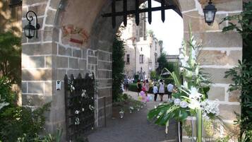 An arched stone gateway with a wooden fence and decorative plants, leading to a building with a stone facade.