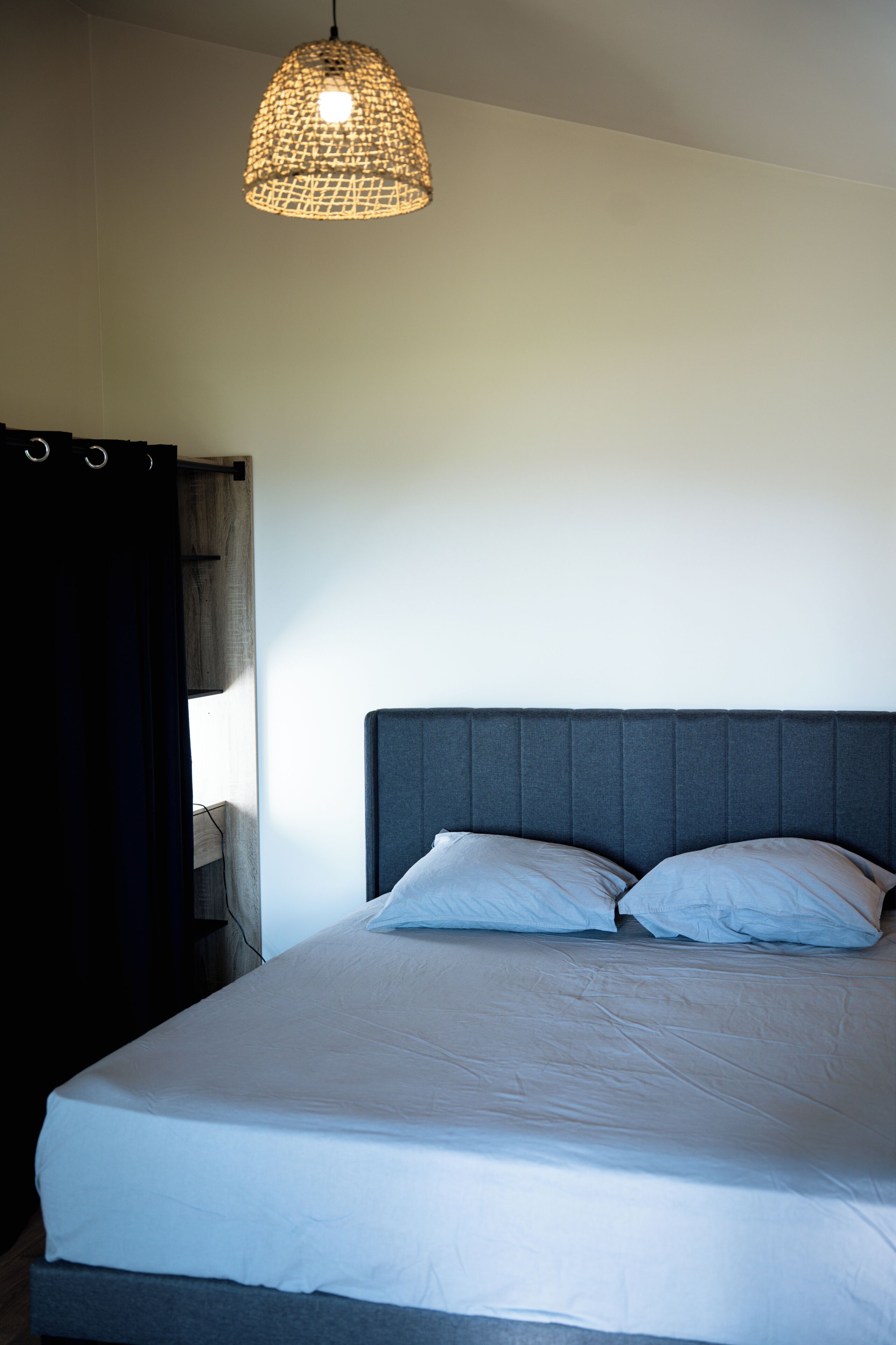 Ein Bett mit grauem Kopfteil, weißer Bettwäsche und einer gewobenen Pendelleuchte.
