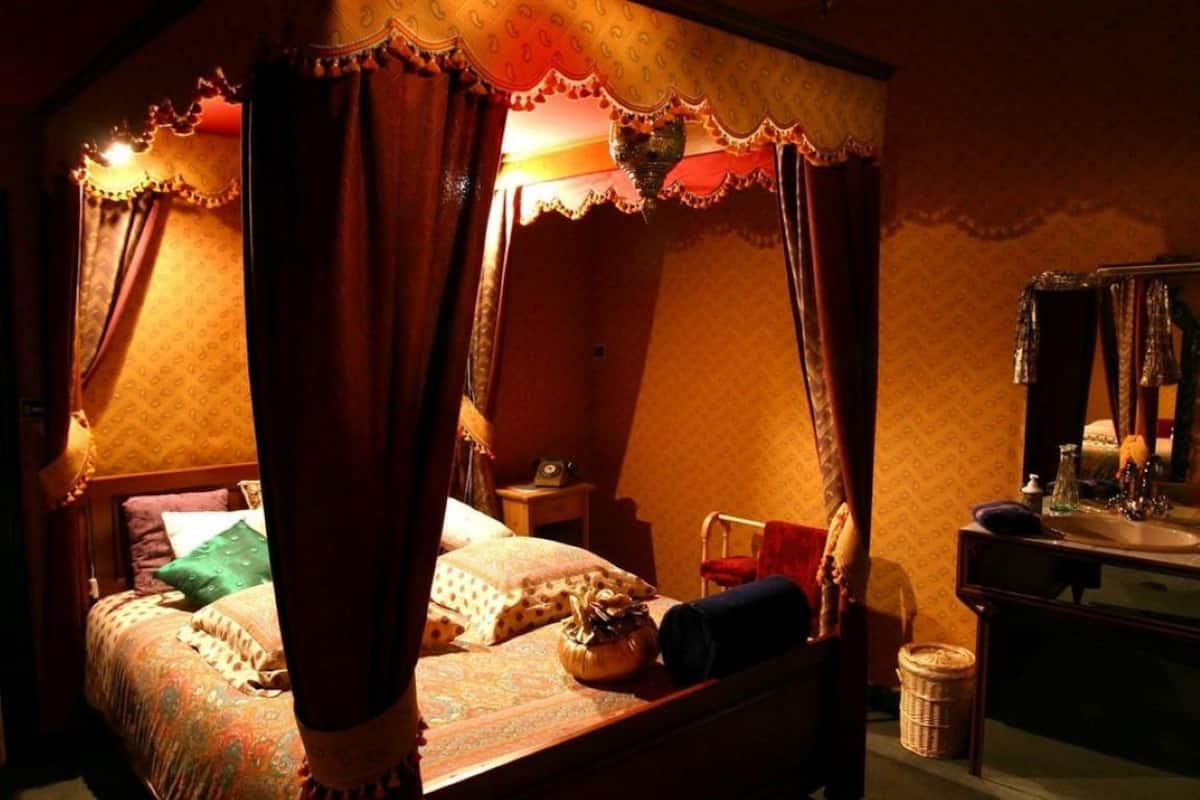 Ein Himmelsbett mit einem gemusterten Überwurf und Kissen, ein Nachttisch mit Lampe sowie ein Spiegel über der Kommode.