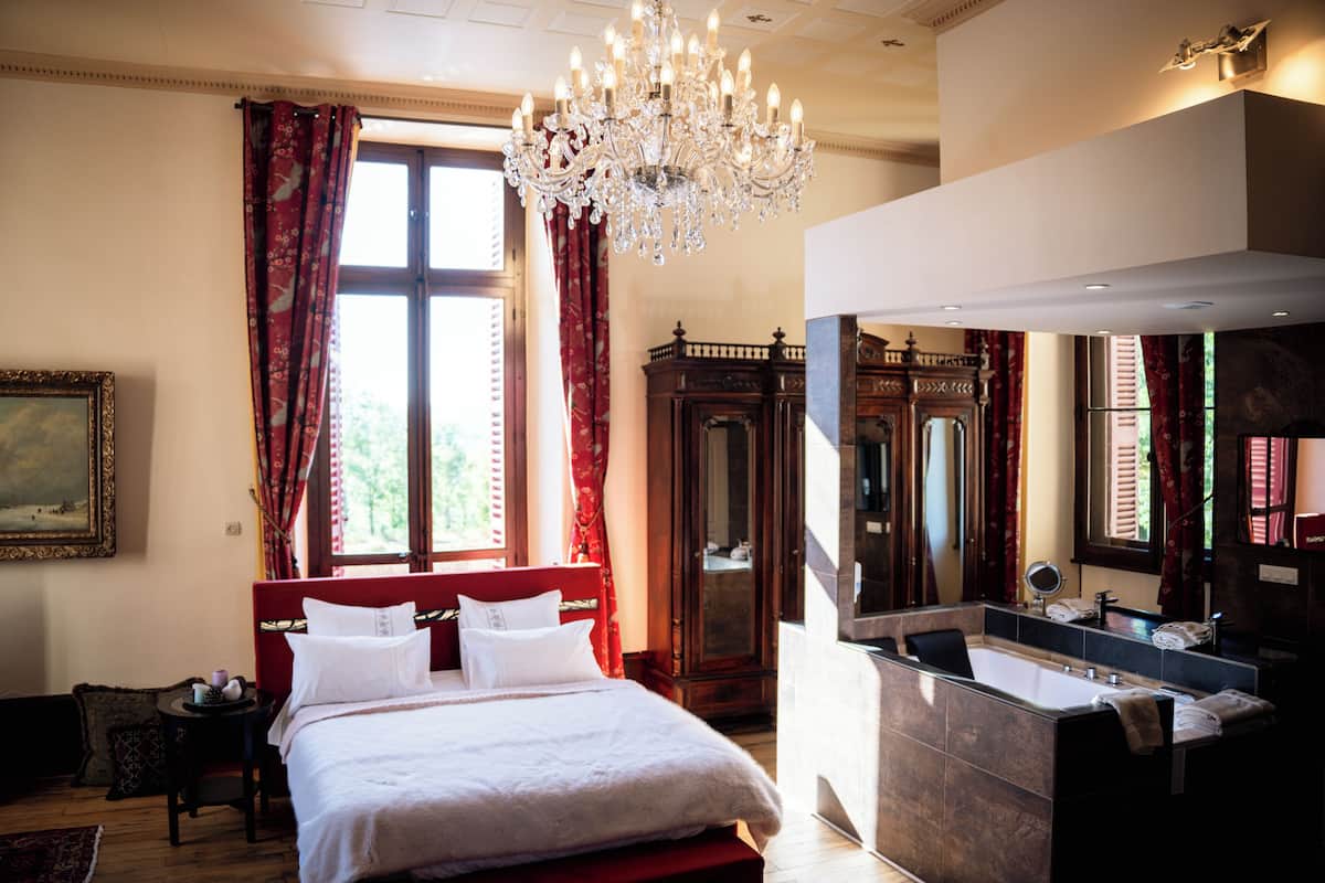 Ein geräumiges Schlafzimmer mit einem großen Bett, einer Kristallleuchtre und Blick ins Freie durch ein Fenster mit roten Vorhängen.