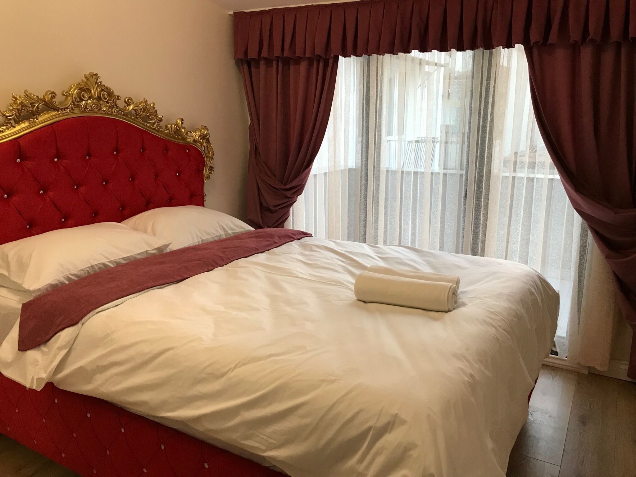 Una cama con cabecera roja y ropa de cama blanca, una toalla color beige y una ventana con cortinas traslúcidas.