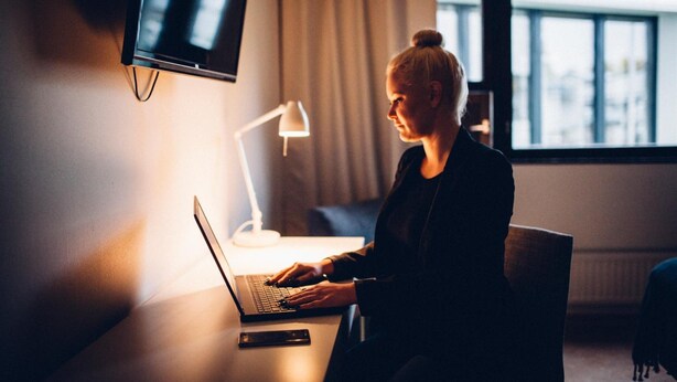 A person using a laptop in a room with a TV mounted on the wall, a lamp on a table, and a window with curtains. - Hotelli Kittilä (Kittila)