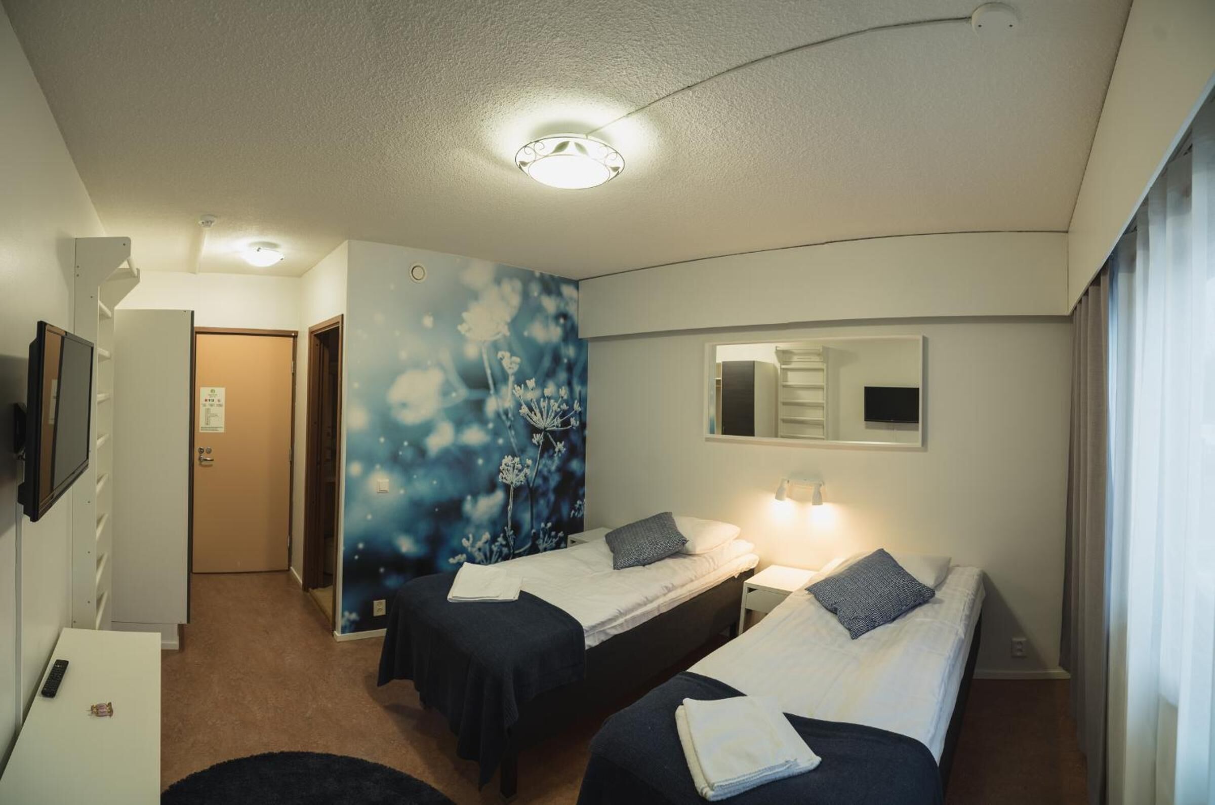 A hotel room with two single beds, a desk, a chair, a TV, and a wall with a floral mural.