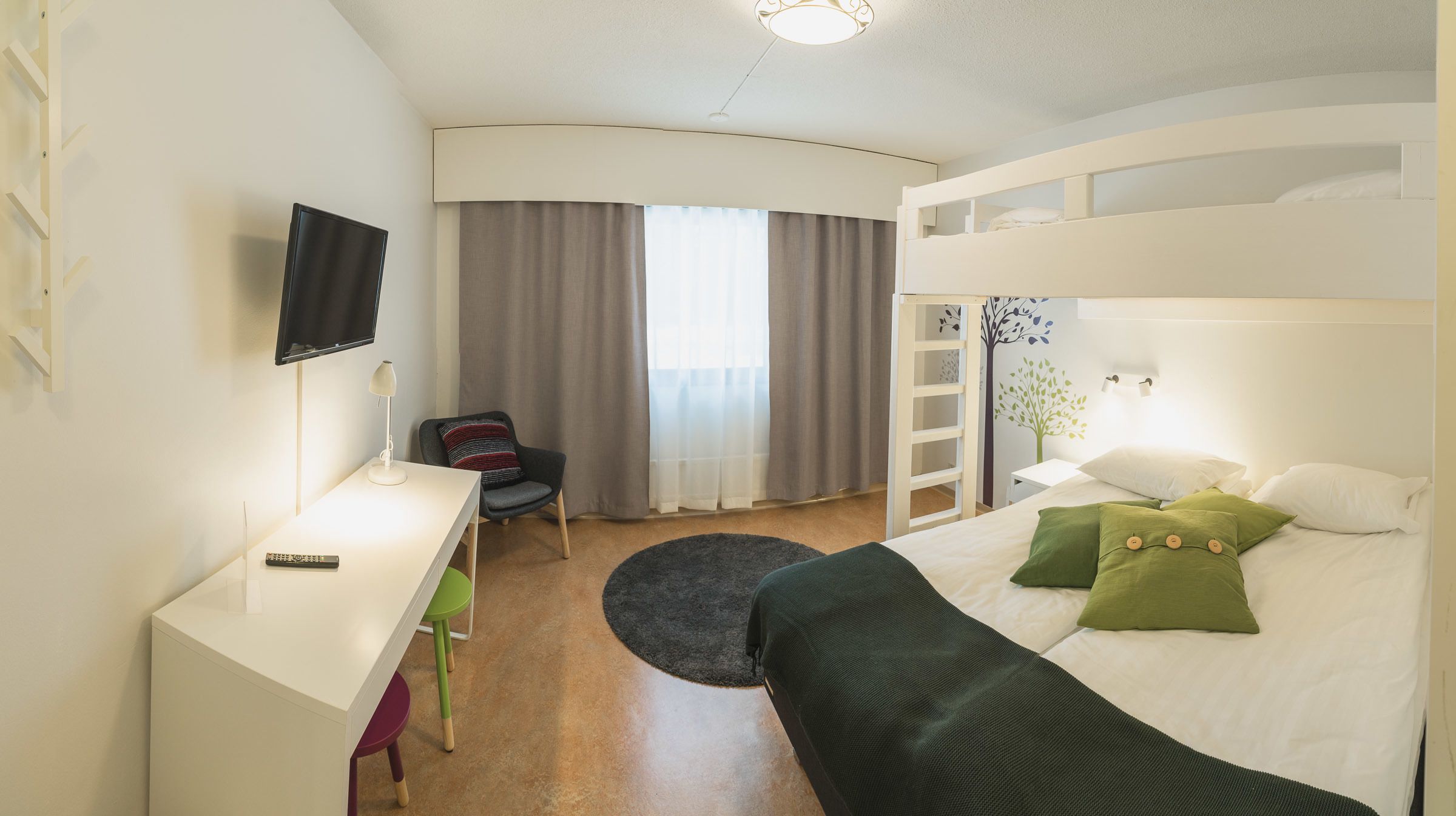 A hotel room with a bed, a desk with a TV, a chair, a window with curtains, and a wall with a tree decal.