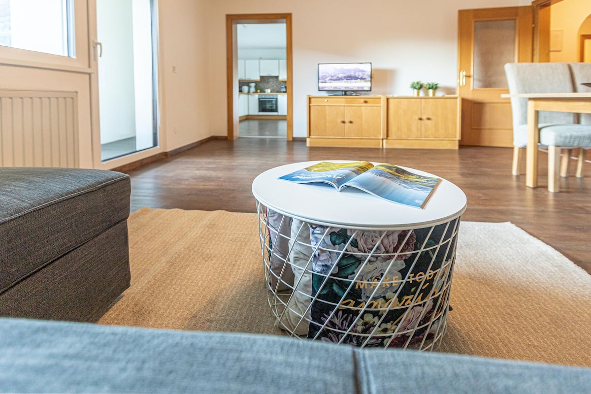 A living room with a grey sofa, a round coffee table with magazines, a wooden cabinet, and a television.