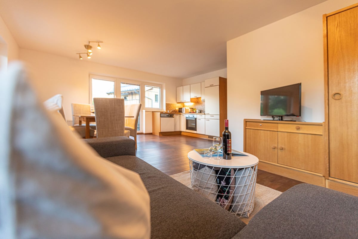 A modern living room with a sofa, a round coffee table with a bottle and glasses, a television on a wooden cabinet, and a kitchen in the background.