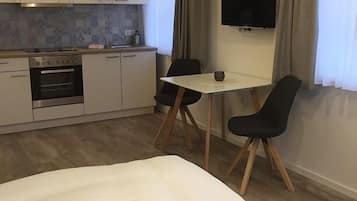 A compact hotel room with a bed, a small kitchenette, a dining table with two chairs, and a TV mounted on the wall.