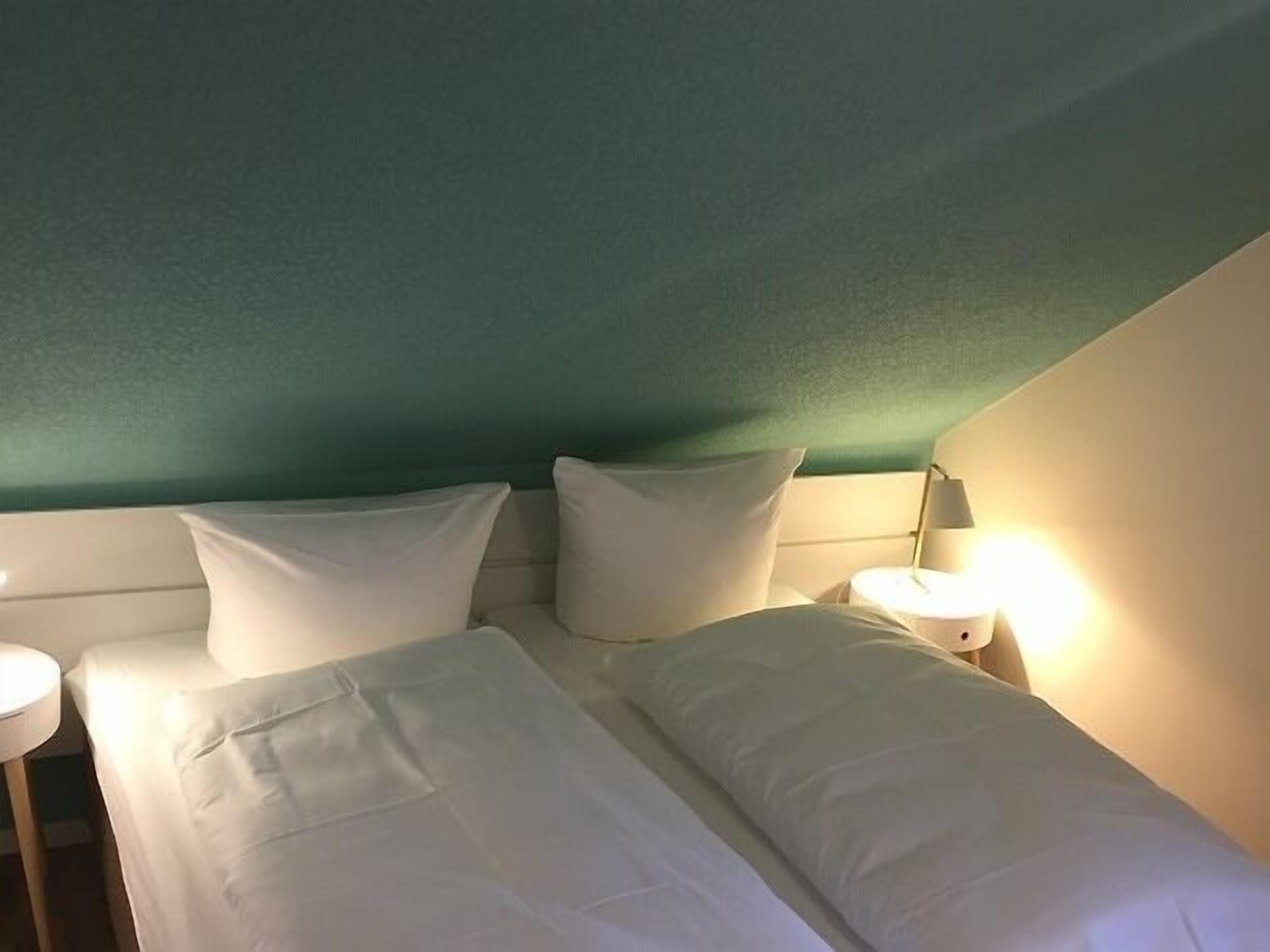 Cama doble con ropa de cama blanca en una habitación con paredes verde azulado y cabecera blanca.