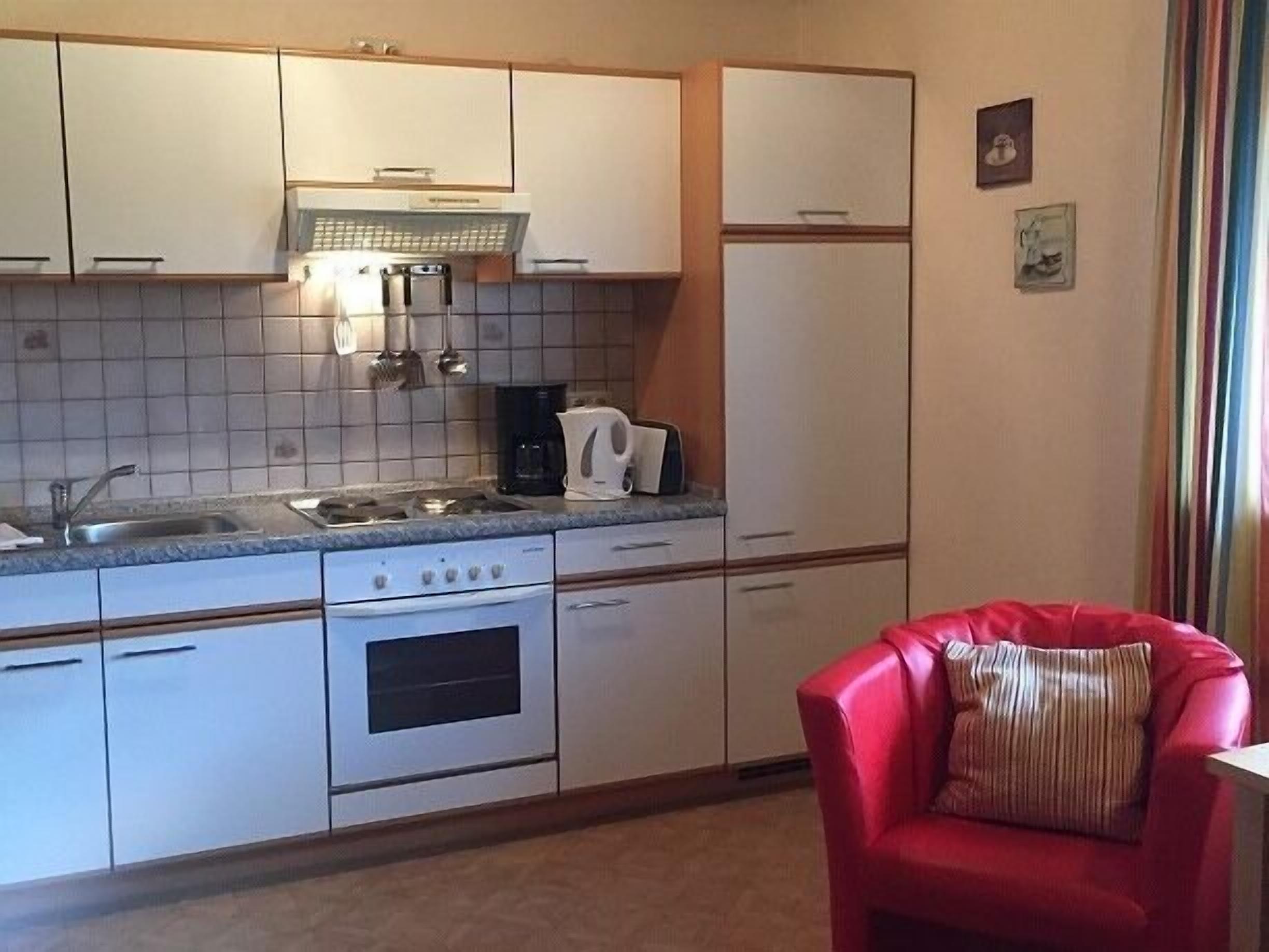 A compact kitchen with white cabinets, a stove, a coffee maker, and a red chair with a striped pillow.