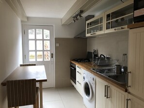 A compact kitchen with a washing machine, a dining table, and a window with multiple panes. - Marble Apartments Tooting Broadway (London)