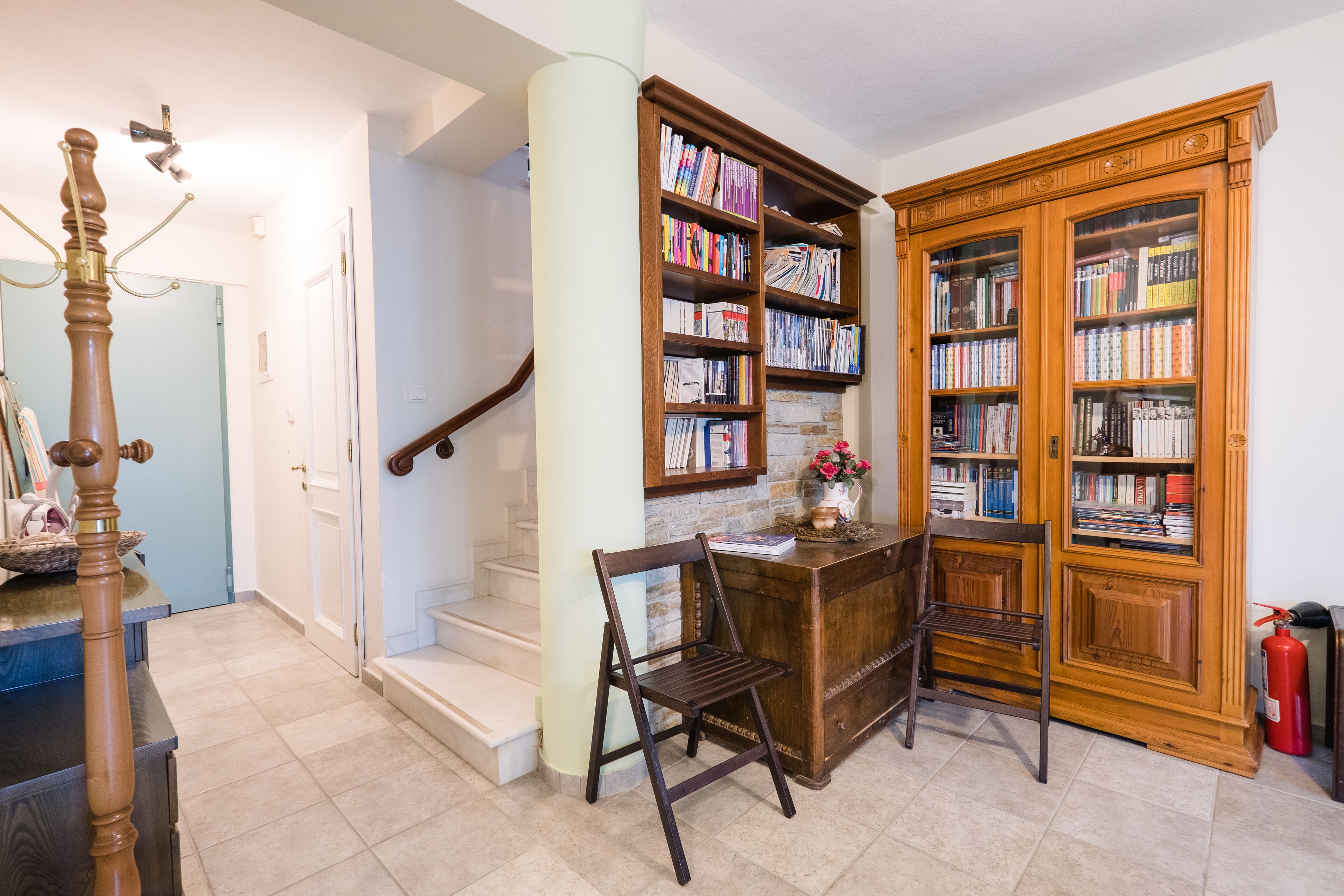 A library with bookshelves, a wooden desk, two chairs, a fireplace, and a staircase.