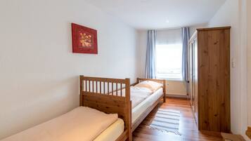 A small, well-lit room with two wooden beds, a wooden wardrobe, and a window with curtains.