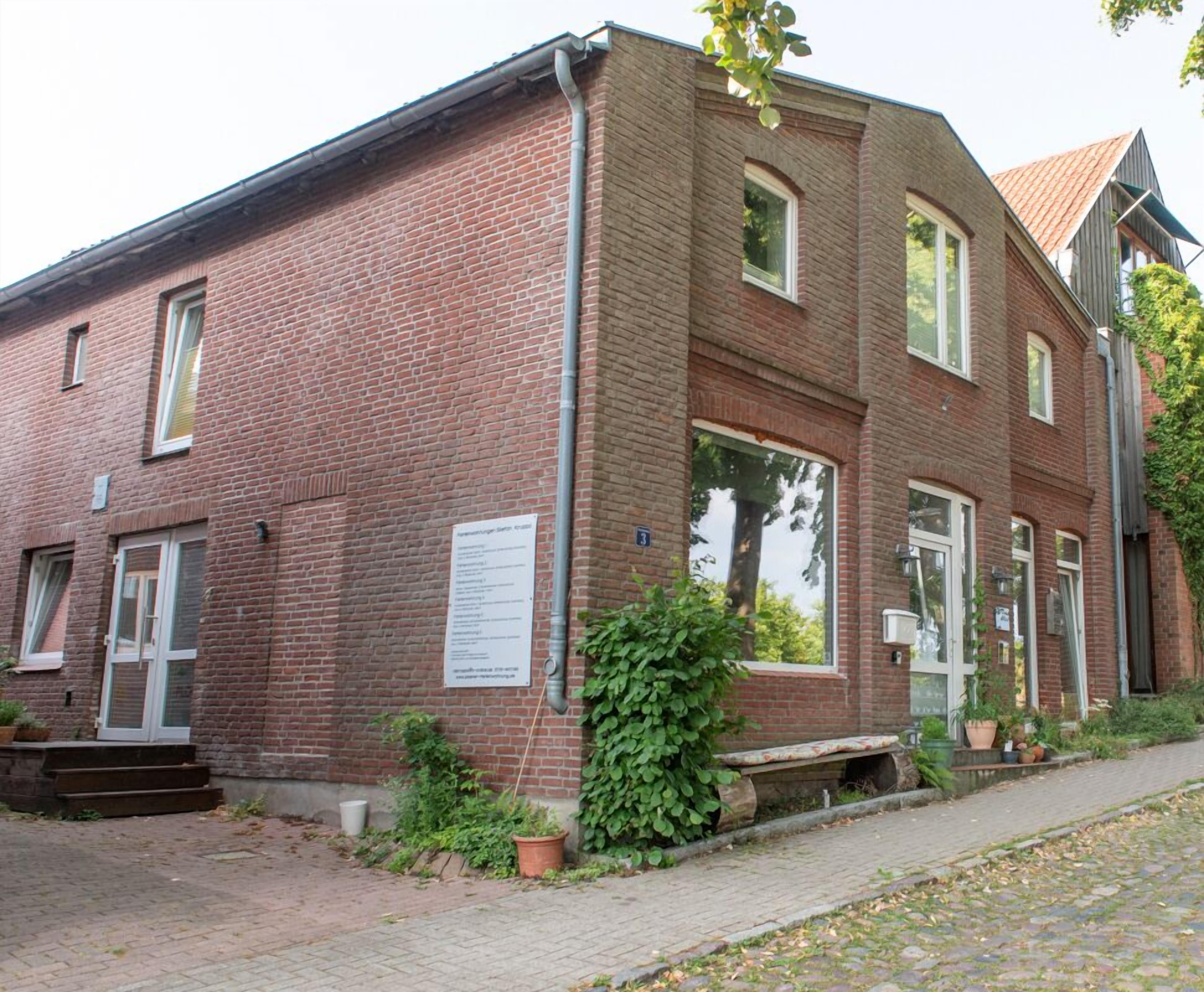 Een gebouw van rode bakstenen met een witte deur, grote ramen en een klein tuintje met plantenpotten.