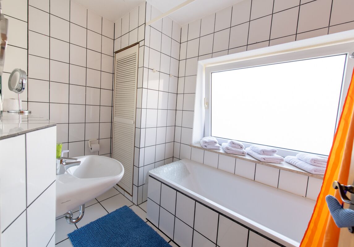 Een badkamer met witte betegelde wanden, een wastafel, een badkuip en een raam met handdoeken op de vensterbank.