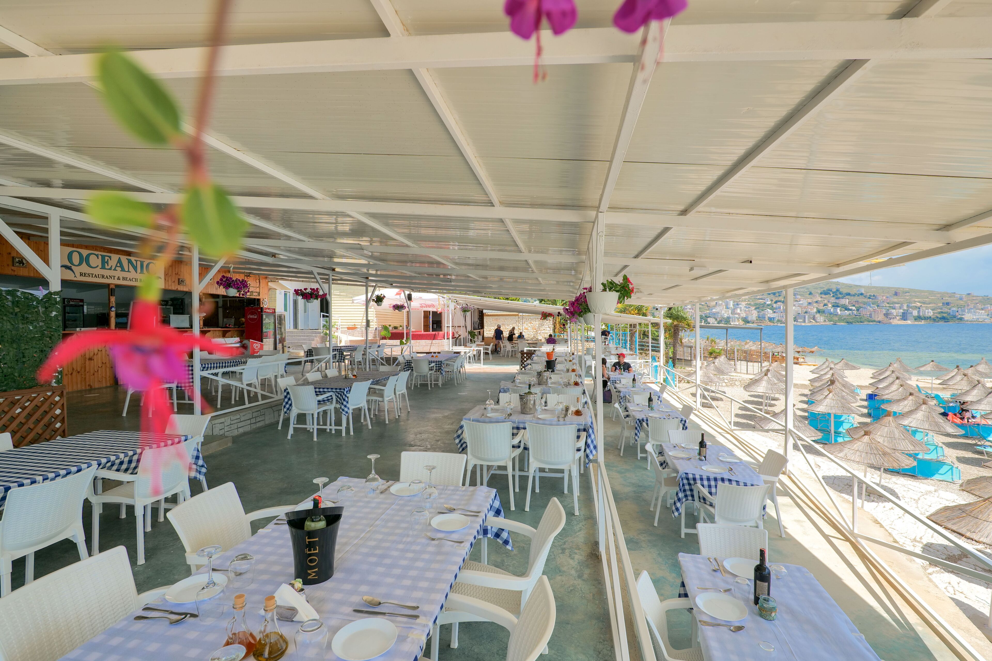 Un espacio exterior para comer con mesas y sillas, vista a la playa y el restaurante "Oceanic" al fondo.