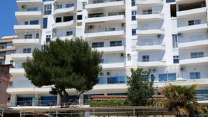 Un edificio blanco de varios pisos con balcones, unidades de aire acondicionado y un árbol en primer plano.