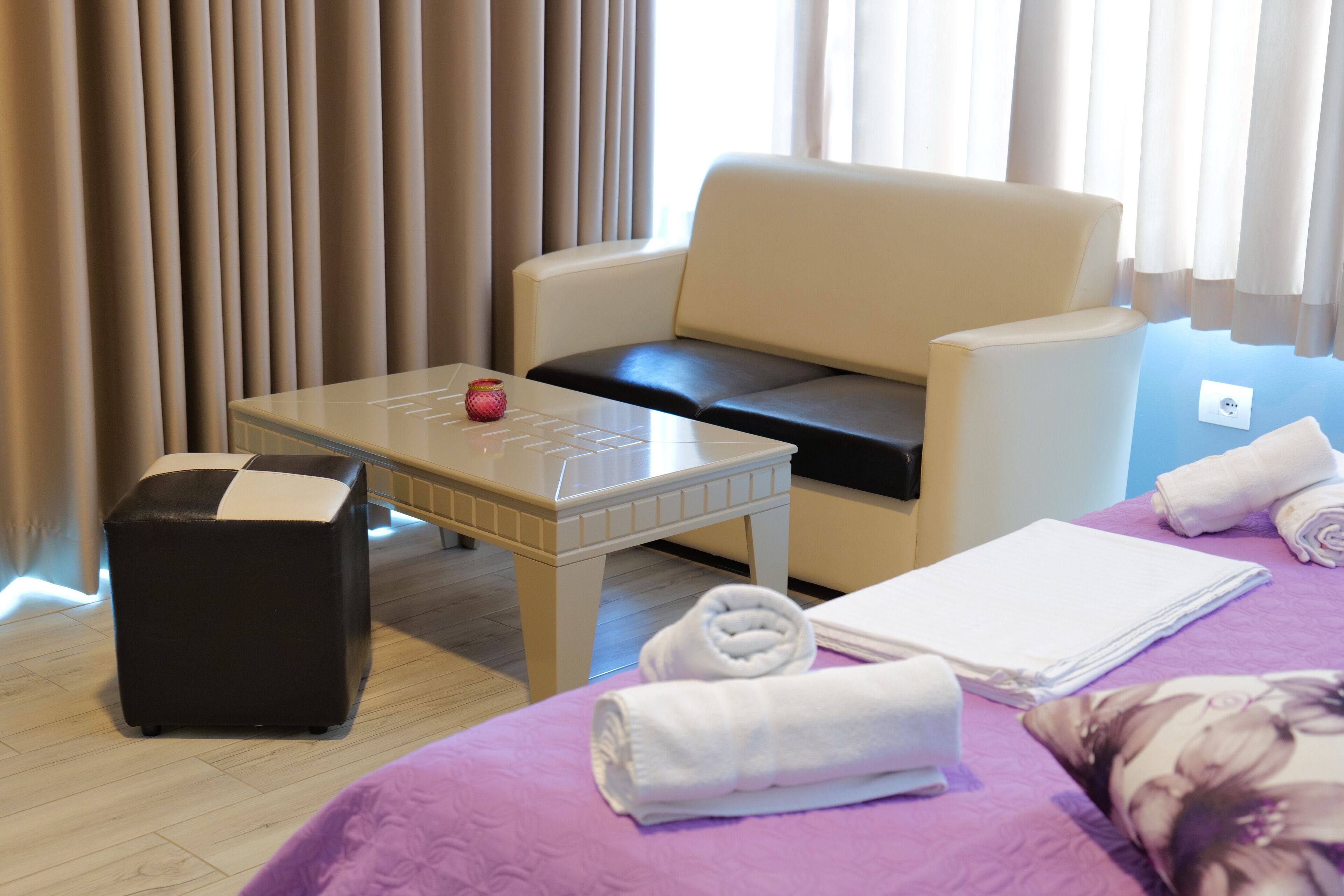 A hotel room with a bed, a sofa, a small table with a red object, a black ottoman, and towels.