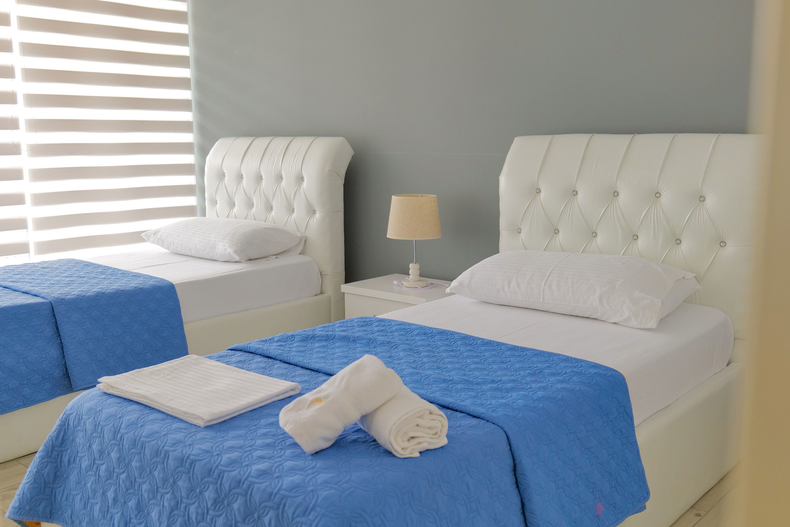 A hotel room with two beds, each with a blue bedspread and white pillows. There is a nightstand with a lamp between the beds.