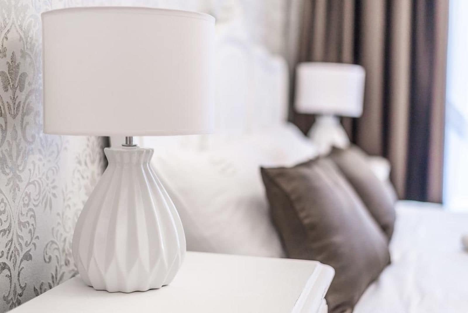 A white bedside table with a white lamp, a bed with white and brown pillows, and a patterned wallpaper in the background.