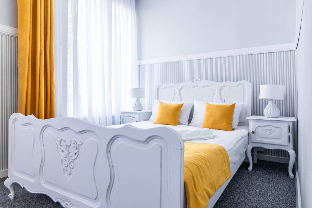 A white four-poster bed with a yellow throw blanket, two bedside tables with lamps, and a window with yellow curtains.
