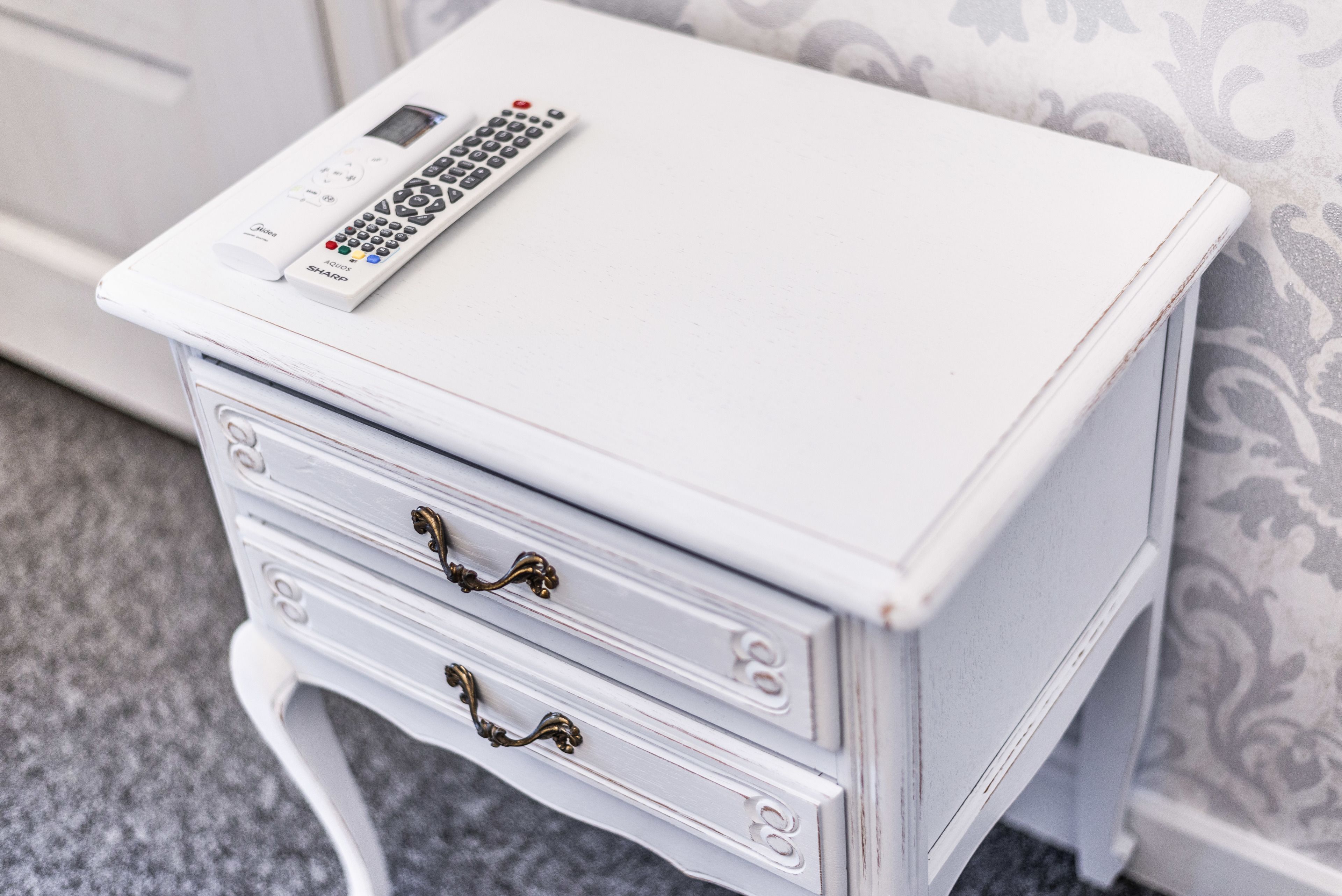 A white bedside table with a remote control, two drawers with brass handles, and a patterned wallpaper background.