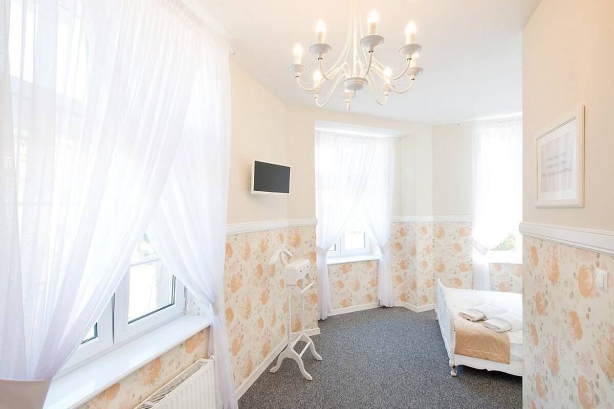 A bedroom with a bed, a window with curtains, a radiator, and a chandelier.