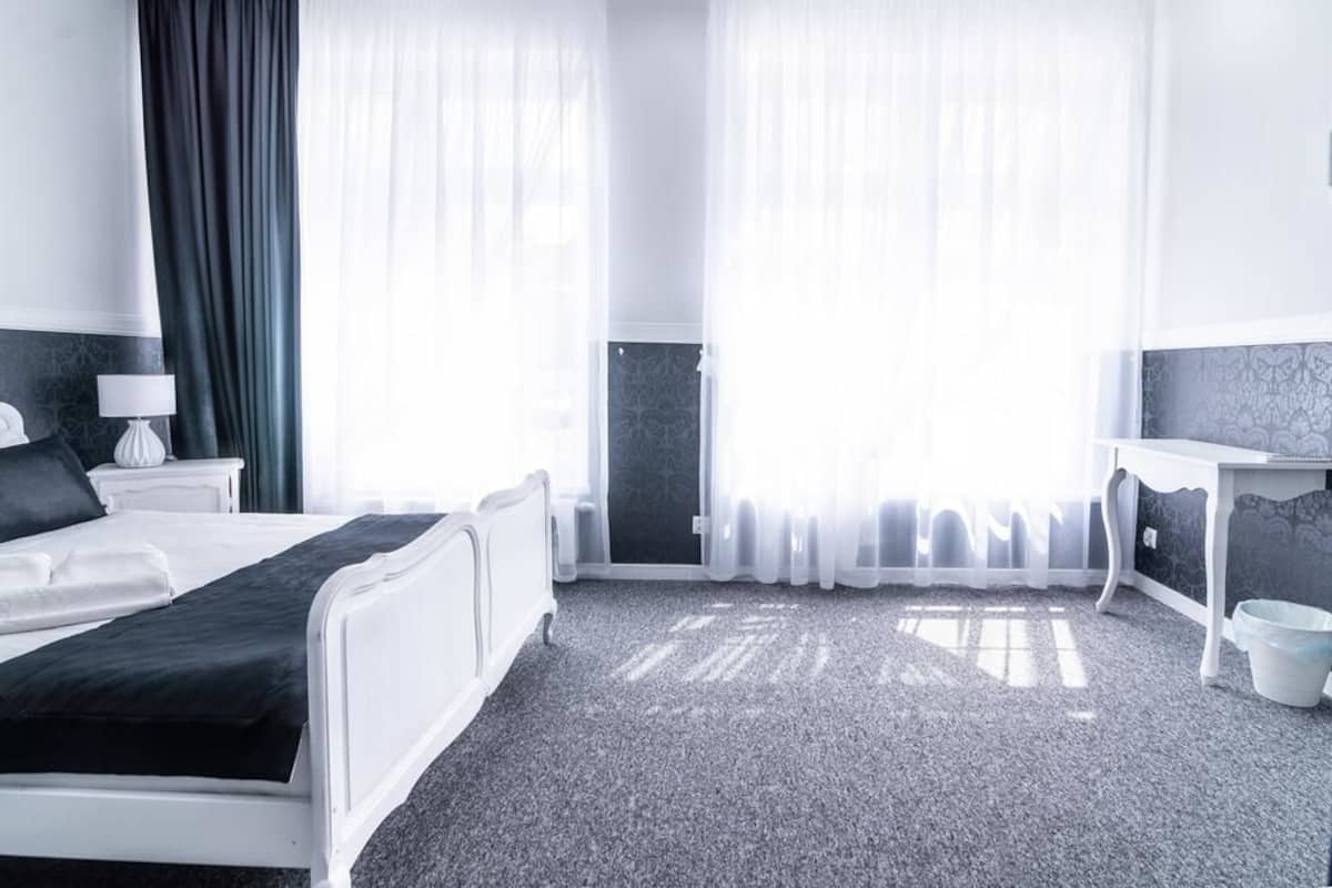 A hotel room with a bed, a desk, and a chair. There is a window with curtains and a door with a patterned wallpaper.