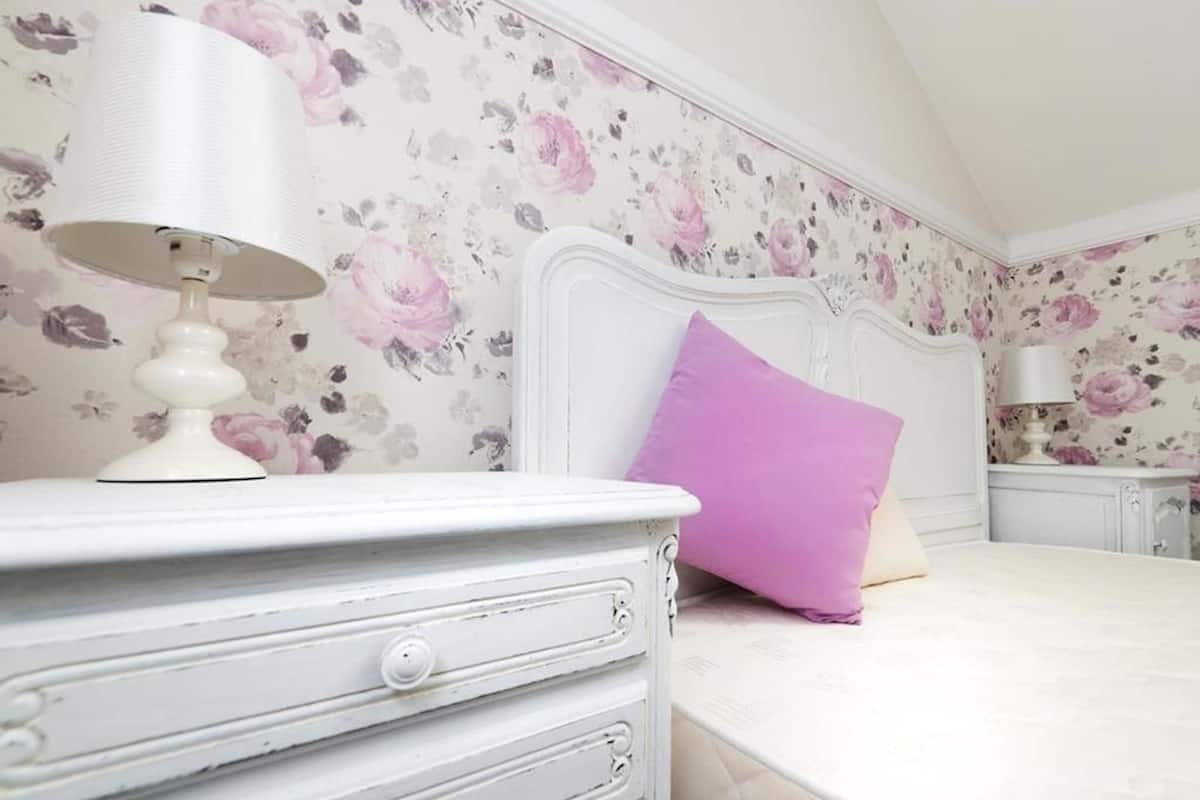 A bedroom with a floral wallpaper, a white headboard, a purple pillow, and a white bedside table with a lamp.