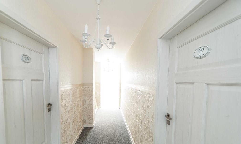 A narrow hallway with white doors, each marked with a number. A chandelier hangs from the ceiling, and the walls have a patterned wallpaper.